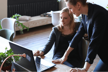 Two female colleagues are helping each other on a laptop at a desk.