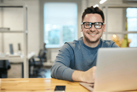 A man wearing glasses and a blue jumper smiling while using a laptop