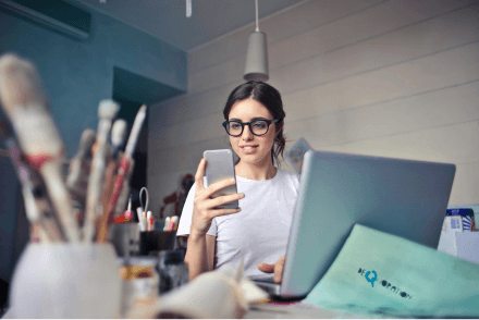 A woman wearing glasses is sat using both her smartphone and laptop. She is surrounded by paint and paintbrushes.