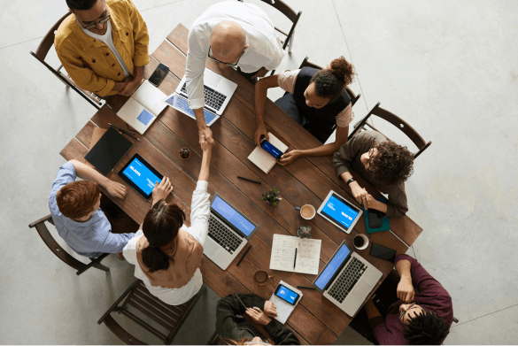 A team of 8 people are sat around a table using laptops, shaking hands and working together. The image is taken from above.