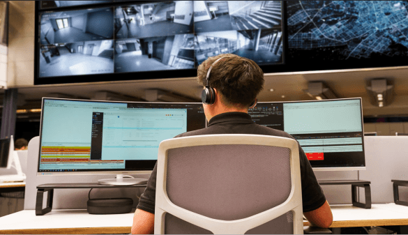 A man sat at a desk wearing headphones, operating multiple screens and watching CCTV